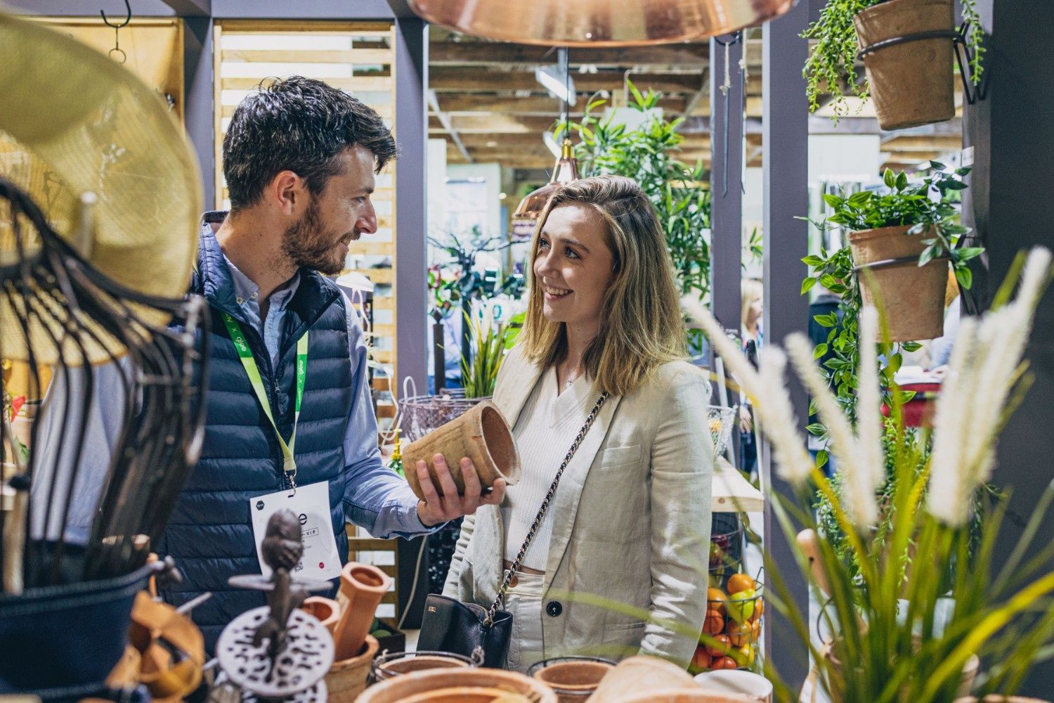 A man in a navy blue gilet holding a plant pot and a woman in a white blazer looking at each other and smiling