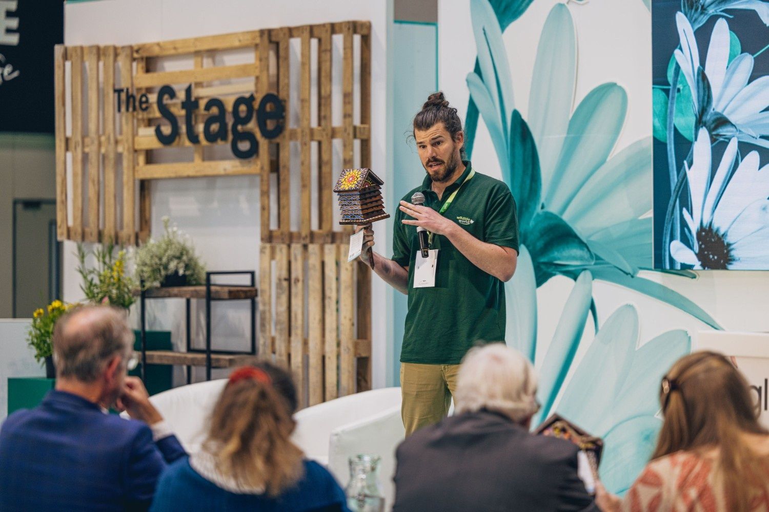 A man wearing a forest green polo shirt is presenting with a microphone whilst holding a small wooden bug house