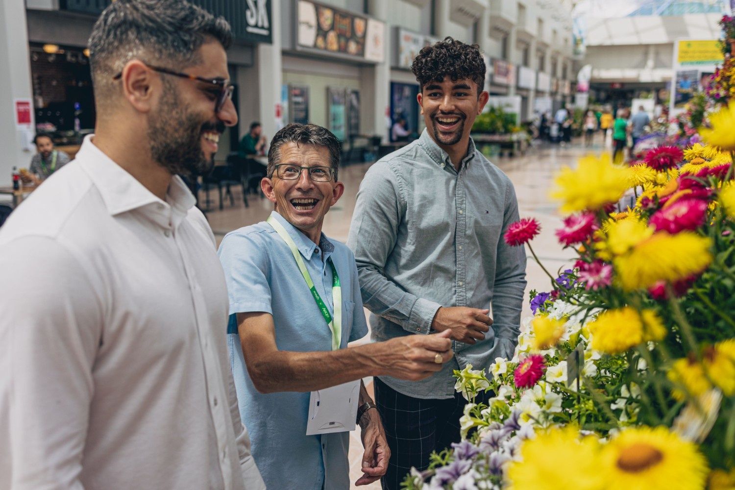 Three men of various ages laughing next to a stall full of vibrant pink and yellow flowers