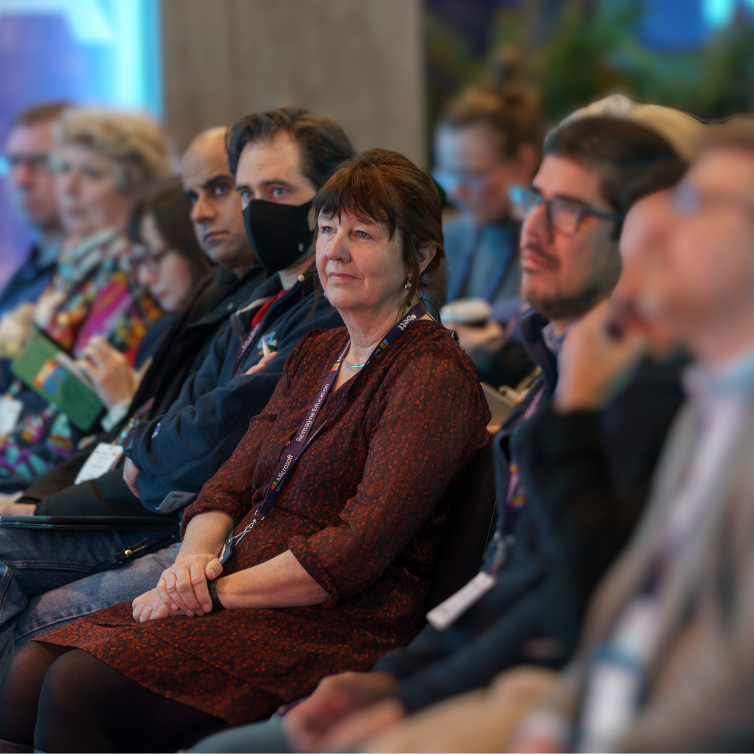 Photo of audience sat down and Gill Ferrell in focus wearing burgundy dresse