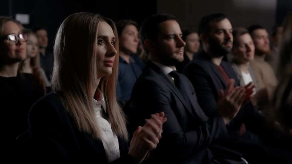 Three professionals in business attire engaged in a discussion, with one person holding a tablet. The background features large glass windows allowing natural light to fill the space.
