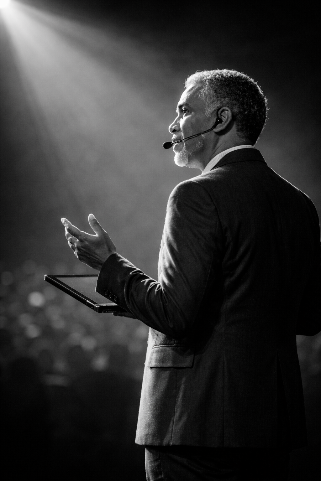 A man in a suit stands on stage, speaking into a microphone with a tablet in one hand. The scene is illuminated by dramatic lighting, creating a captivating atmosphere.