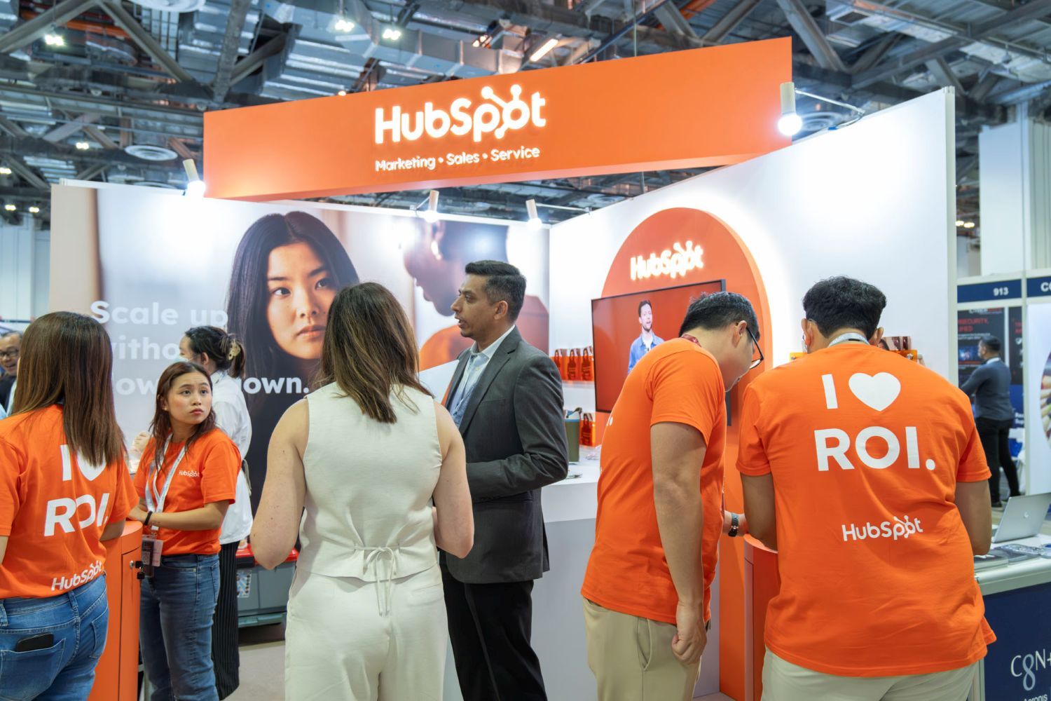 HubSpot exhibition booth at a business trade show, with staff in orange shirts speaking to attendees about marketing, sales, and CRM solutions