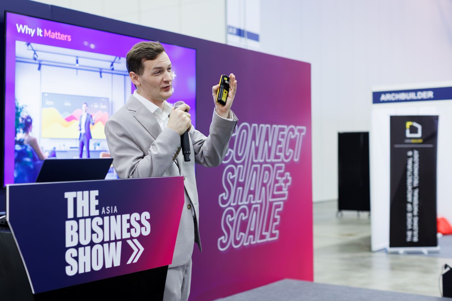 Business conference speaker presenting on stage at The Business Show Asia, holding a device and addressing an audience about growth and innovation