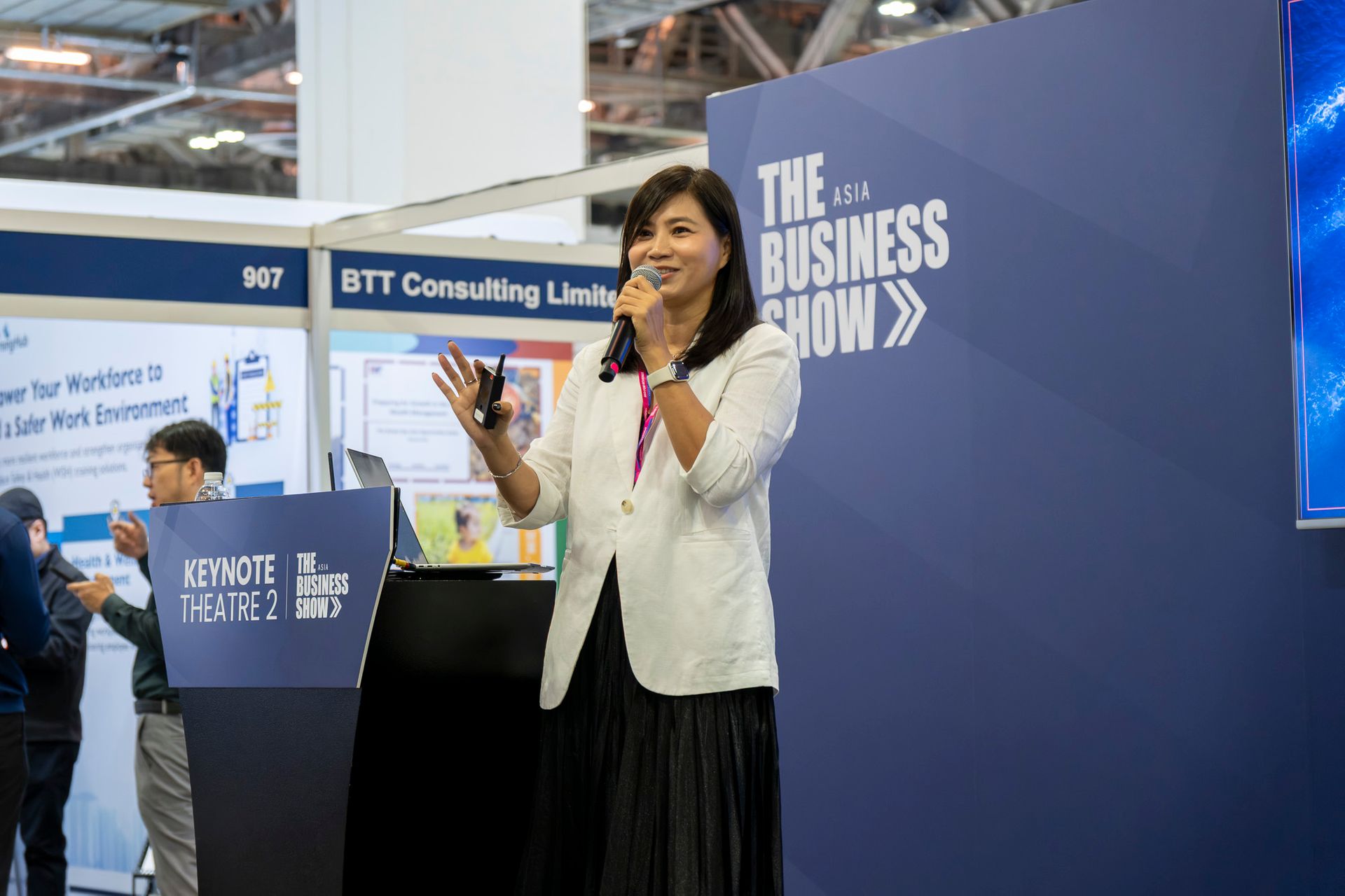 Business conference speaker presenting on stage at The Business Show Asia, holding a device and addressing an audience about growth and innovation