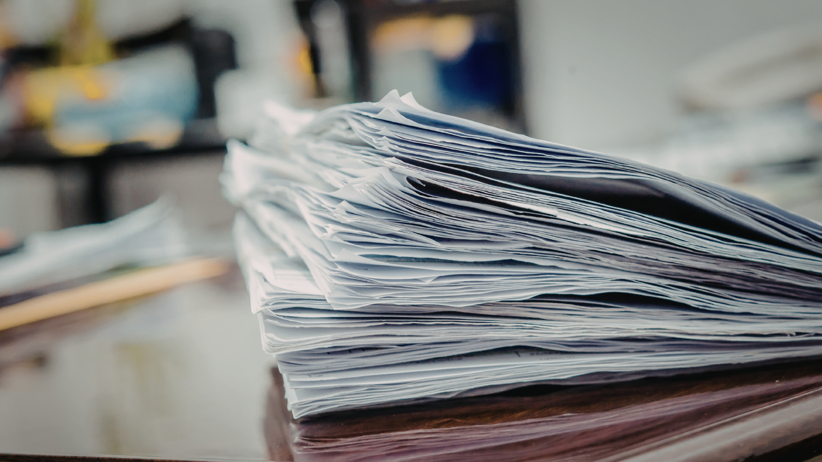 A picture depicting a large stack of documents on a dark wooden desk