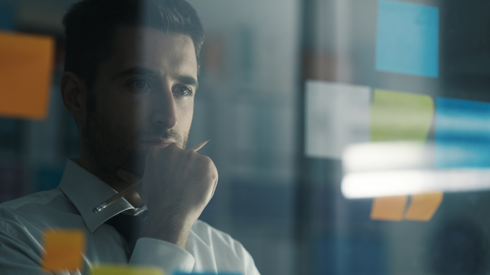 A picture of a man staring at a glass wall covered in post it notes, he is deep in thought