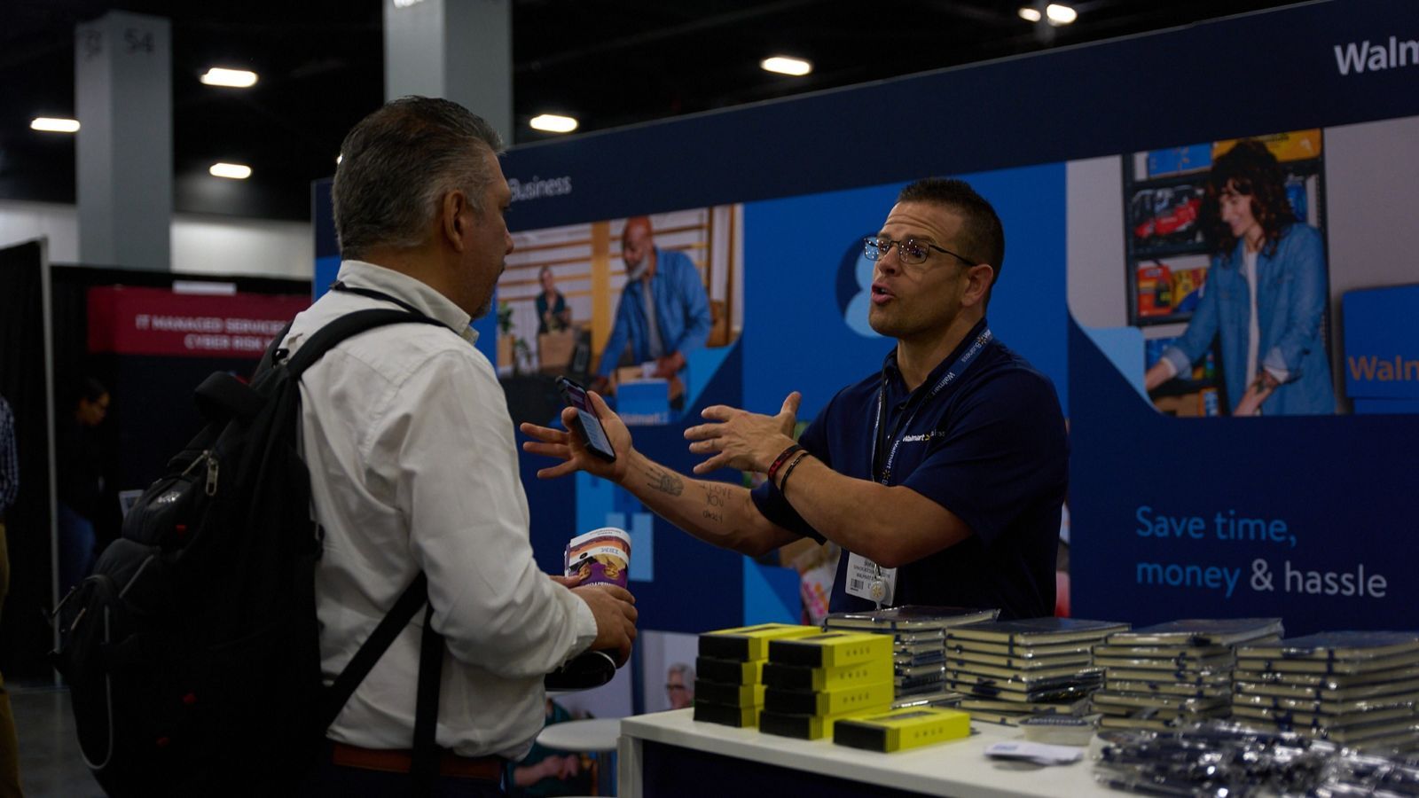Two men talking, one of them is exhibiting to the other at The Business Show Miami, the biggest business expo in the US. The table next to them is full of branded merchandise.