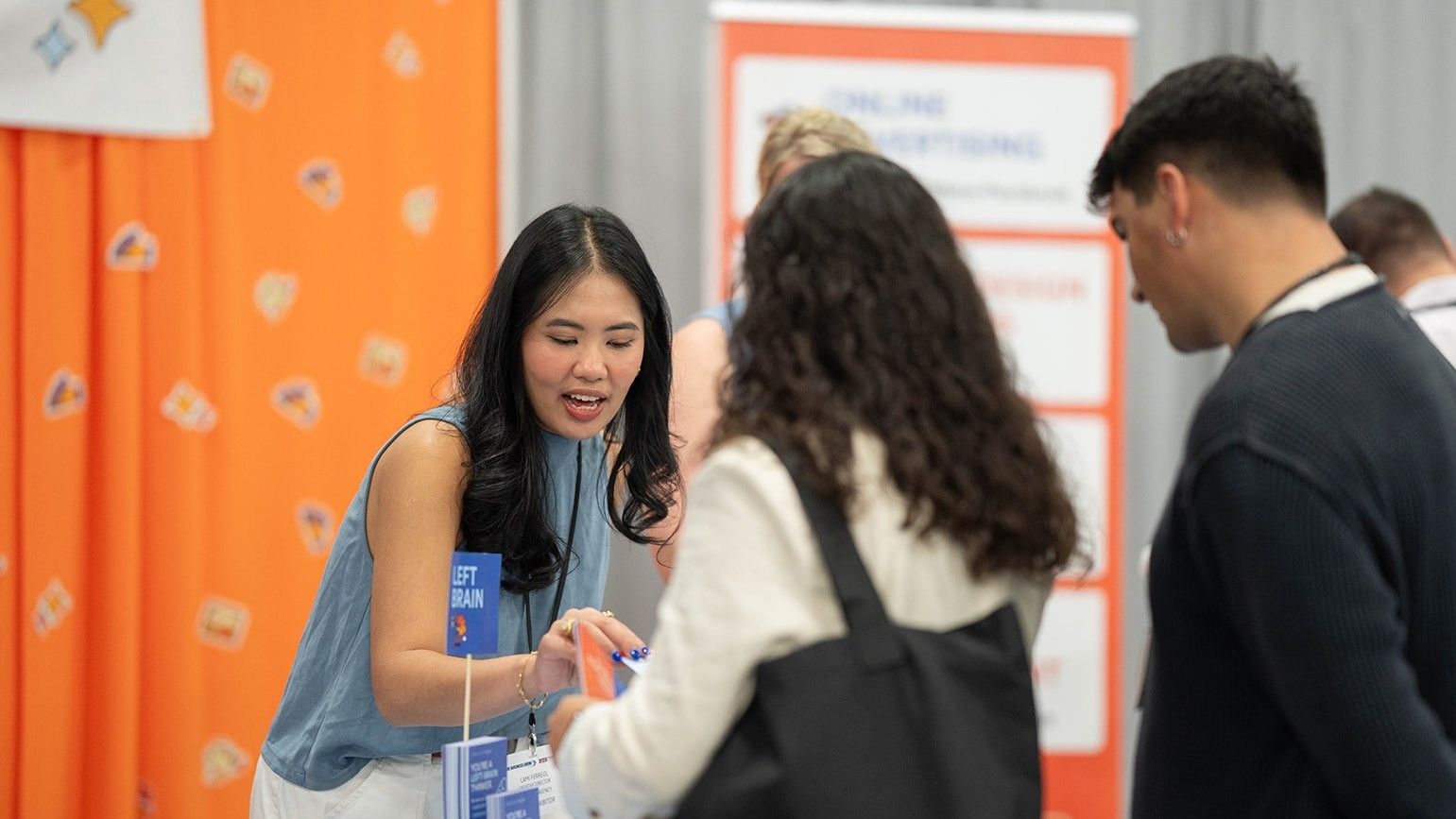 Two people talking. One is an attendee of the show, the other is an exhibitor behind a booth. The booth is clad with orange material