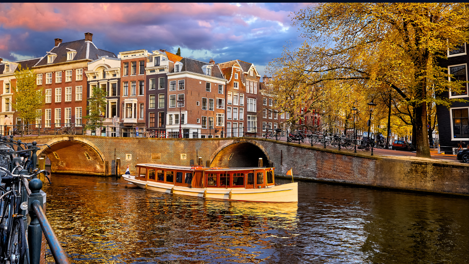 Boat passing under a bridge along Amsterdam’s historic canal lined with autumn trees and canal houses.