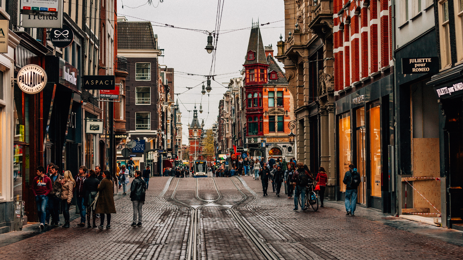 Busy shopping street in central Amsterdam with pedestrians, tram tracks, and historic buildings.