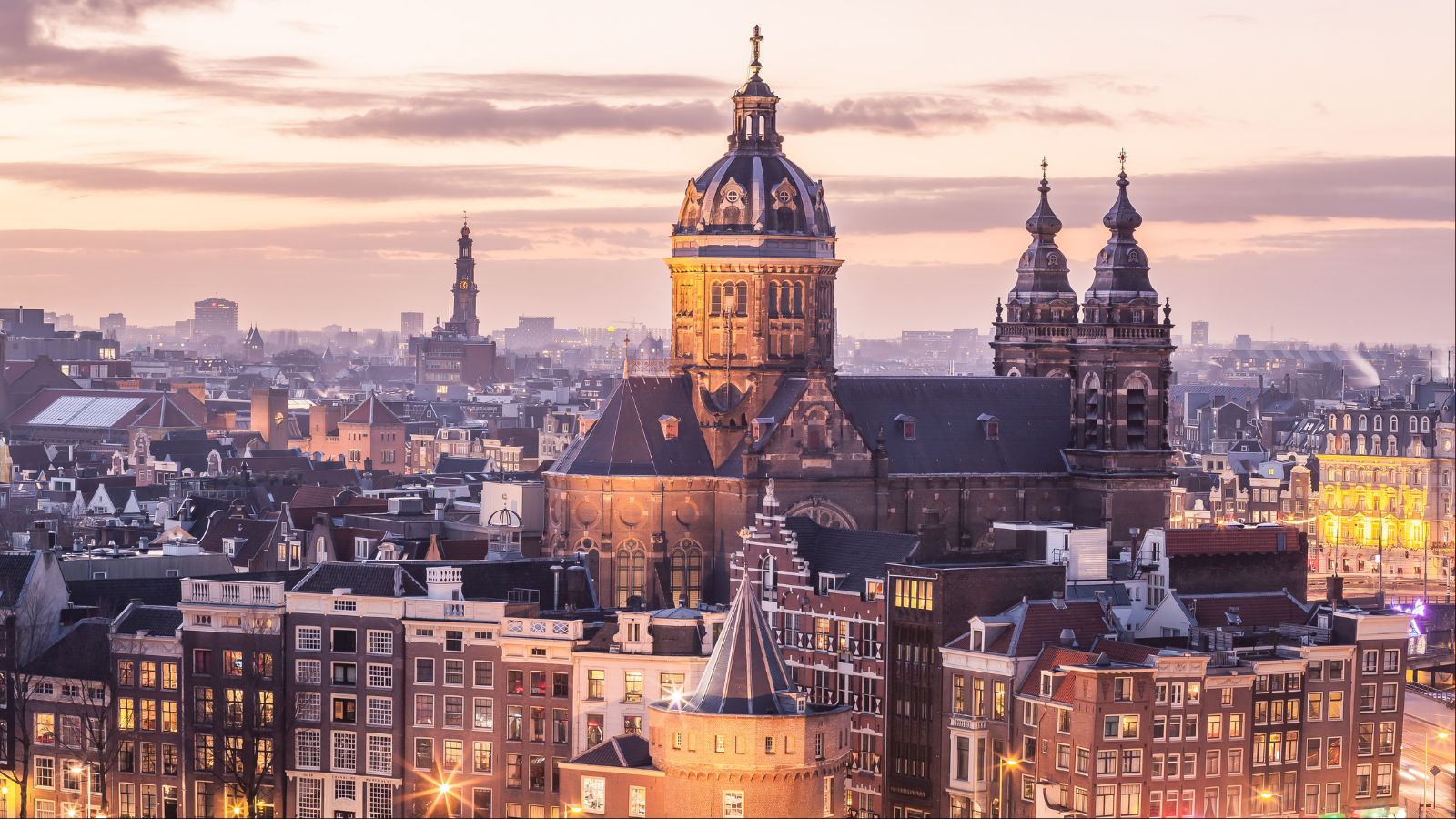 Panoramic Amsterdam skyline at dusk with illuminated buildings, canals, and city lights.