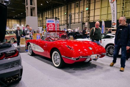 Rear view of a classic red and white Chevrolet Corvette convertible on display at the Classic Motor Show. Attendees walk past the car in a large exhibition hall featuring 