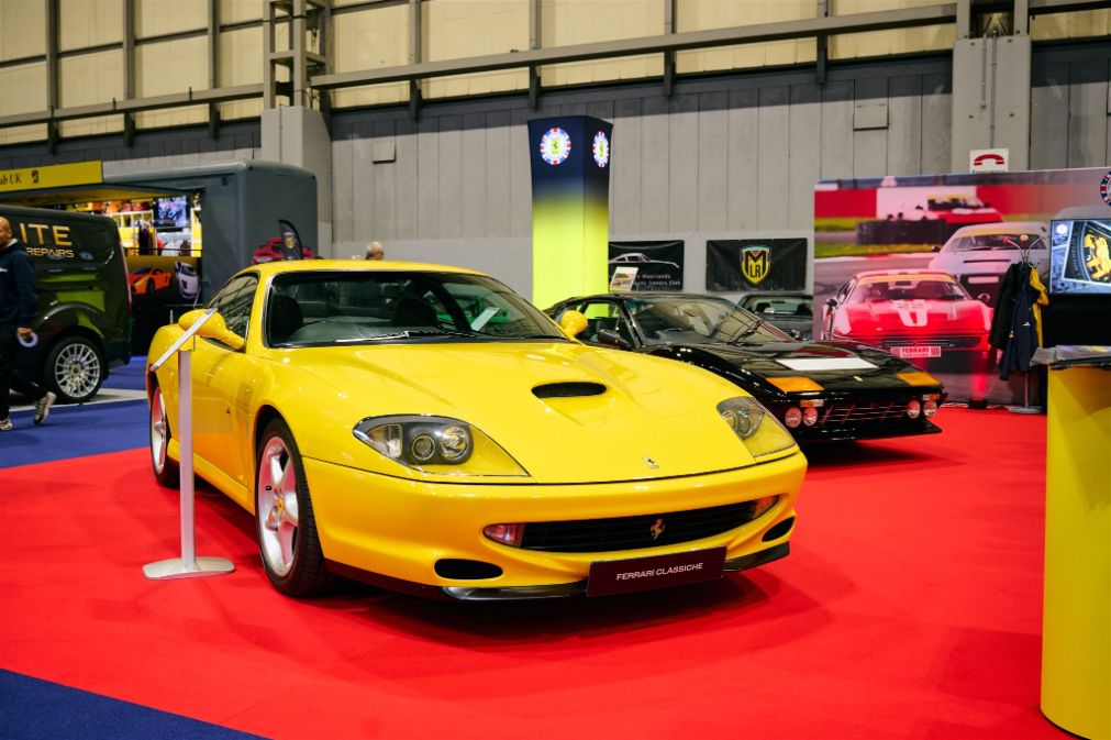 A bright yellow Ferrari 550 Maranello on display at a classic car show, with a black Ferrari Testarossa and other vintage cars visible in the background.