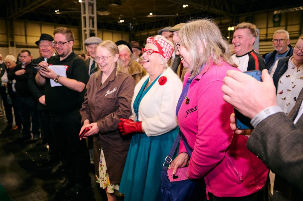 A diverse crowd of classic car enthusiasts, including people dressed in vintage clothing, laughing and smiling at a motor show event.