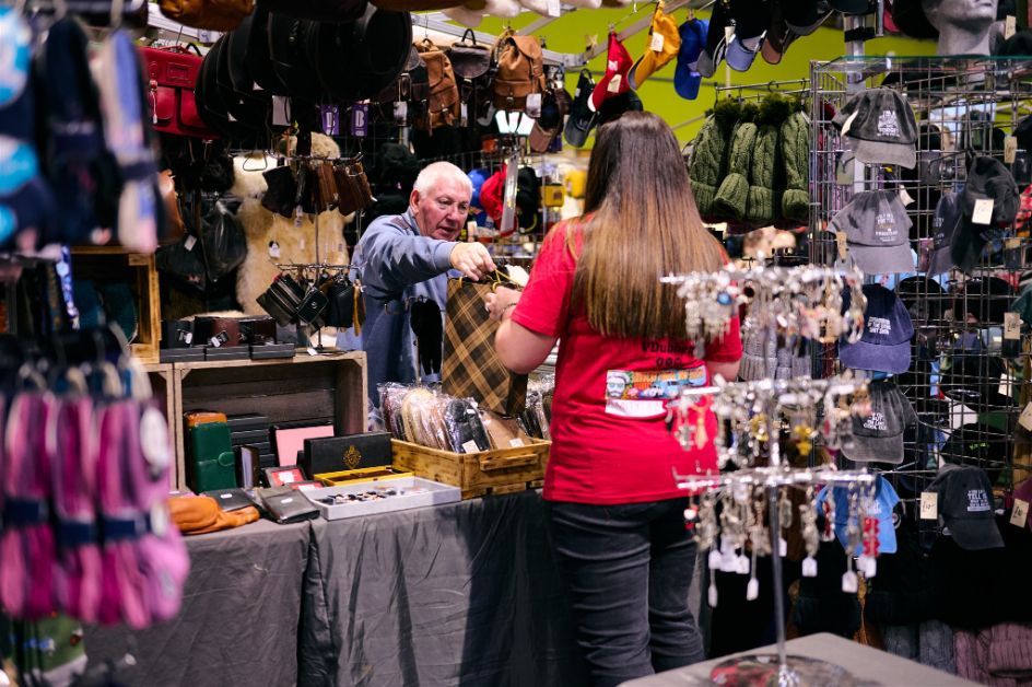 Vendor assisting a customer at a merchandise stall selling classic car themed bags, hats, and accessories at a motor show or enthusiast event.
