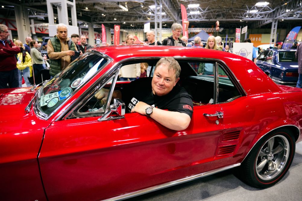 Mike Brewer, host of Wheeler Dealers, sitting inside a classic red Ford Mustang Fastback at a car show.