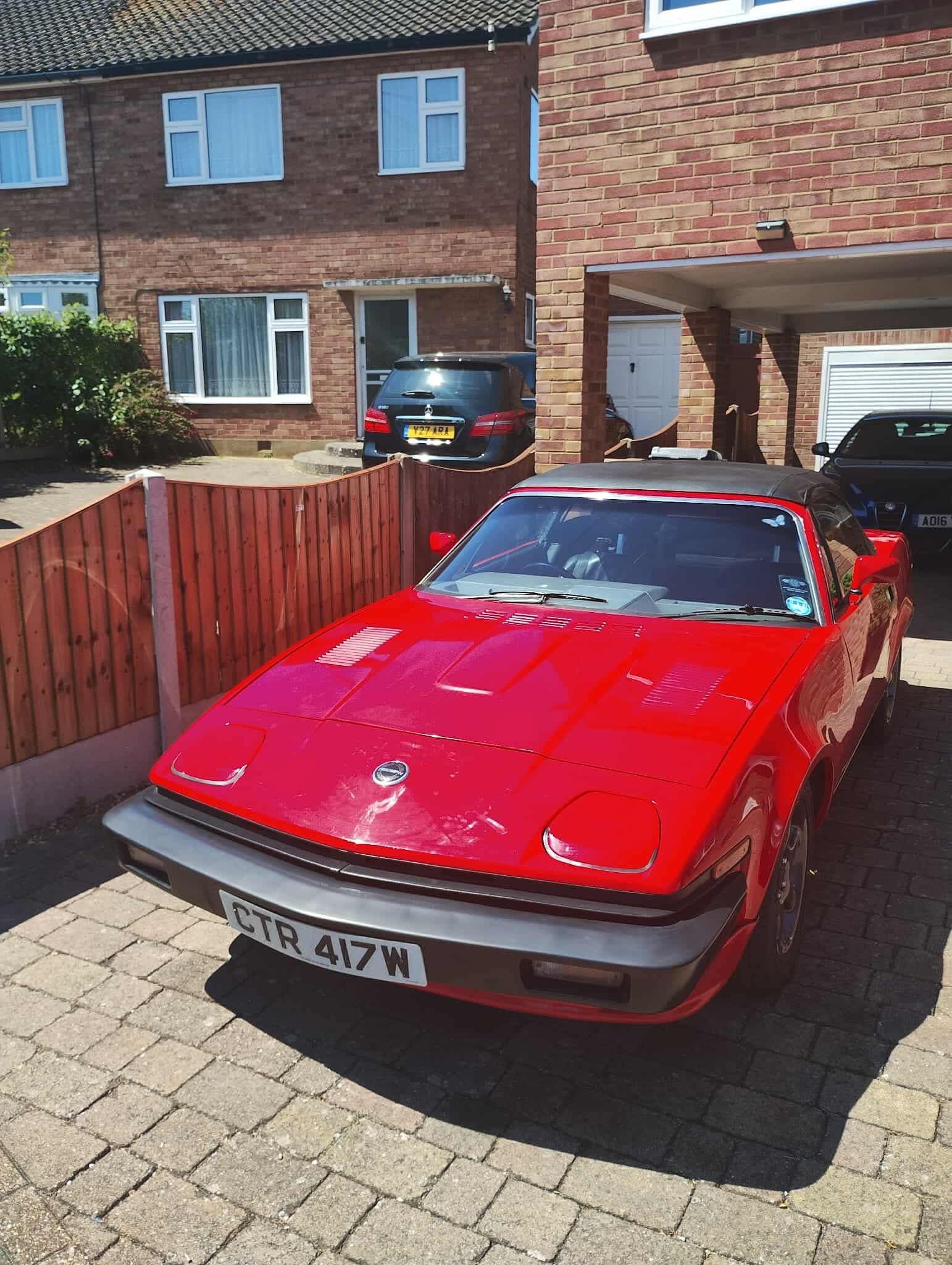 A classic red Triumph TR7 coupe, with a black soft top and license plate "CTR 417W", parked on a brick driveway in front of a residential house.