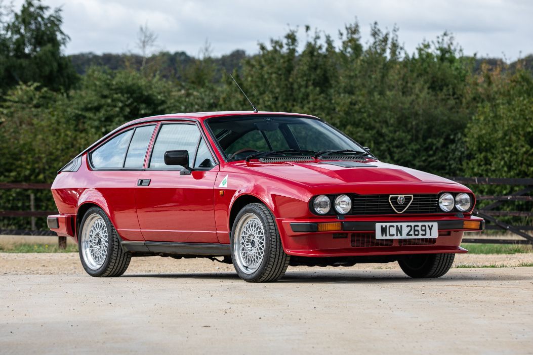 Front-three-quarter view of a classic red 1983 Alfa Romeo GTV6 parked on a paved driveway with trees in the background.