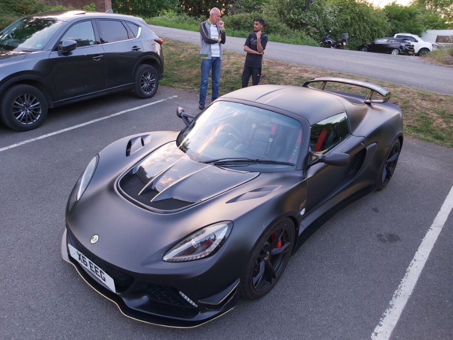 A matte black Lotus Exige sports car with a rear wing parked in a lot at sunset.