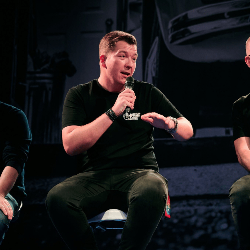 YouTuber Luke from Garage of Luke speaking into a microphone on stage wearing a branded black t-shirt
