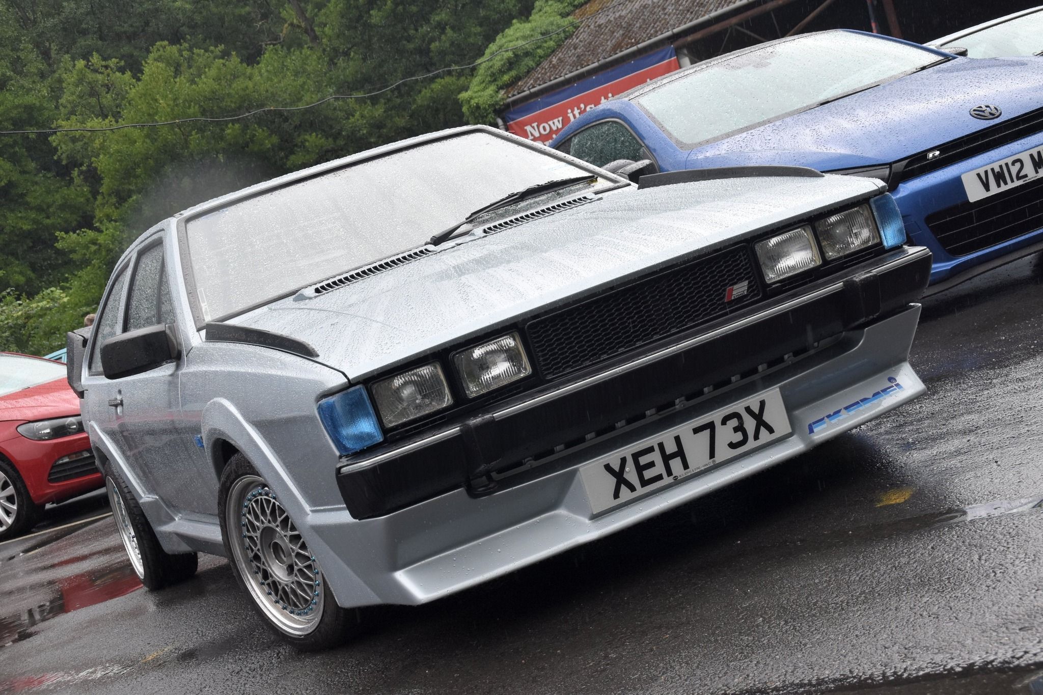 A wet, silver Mk2 Volkswagen Scirocco with a license plate "XEH 73X", featuring custom BBS-style wheels and a front bumper spoiler, parked at a car show on a rainy day.