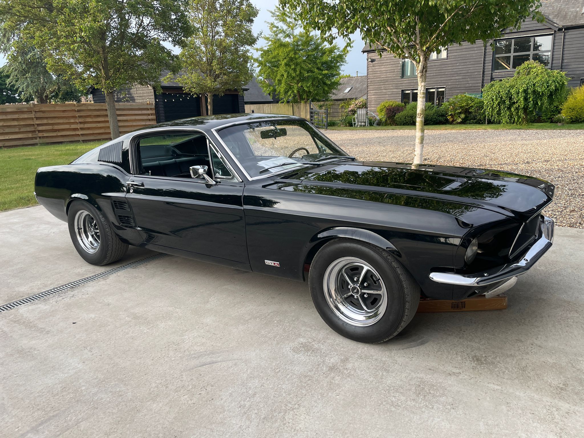 A sleek, black 1968 Ford Mustang Fastback is parked on a concrete driveway in front of a modern, dark-wood-paneled building. The car's polished surface reflects the light, and its classic fastback body style is visible, along with its silver-spoked wheels and a GT badge on the side