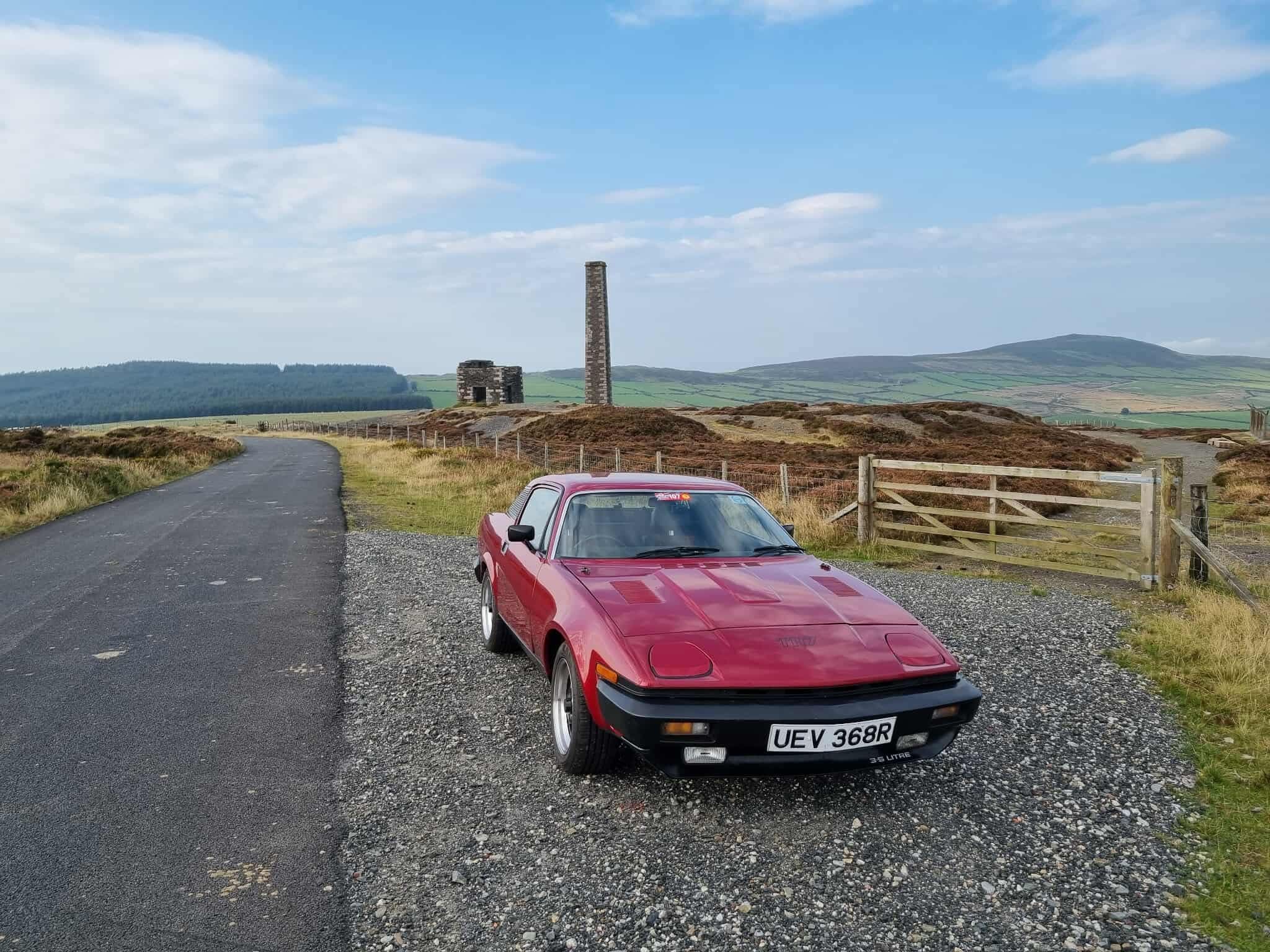 A classic red Triumph TR7 coupe, with license plate "UEV 368R", parked on a gravel shoulder in a hilly, rural landscape with a historic stone ruin and chimney in the background.