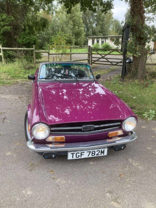 A classic purple Triumph TR6 roadster, with license plate "TGF 782M", parked on a driveway with a black convertible top, a wooden fence, and trees in the background.