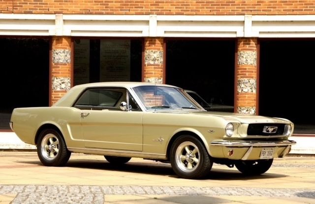 A classic gold 1960s Ford Mustang coupe, owned by Adam Crowton, with aftermarket five-spoke wheels, parked outside a brick building with white pillars.