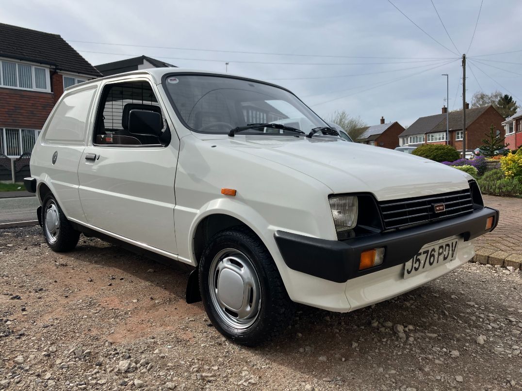 A clean, white classic Austin Metro Van, owned by Alex Mills, with black plastic trim and original style wheels, parked on a gravel driveway.