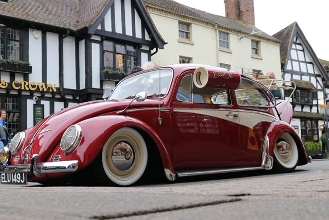 A heavily customized, slammed red and cream Volkswagen Beetle with whitewall tires and a roof scoop/horn, owned by Andy Green, parked in a historic UK town center.