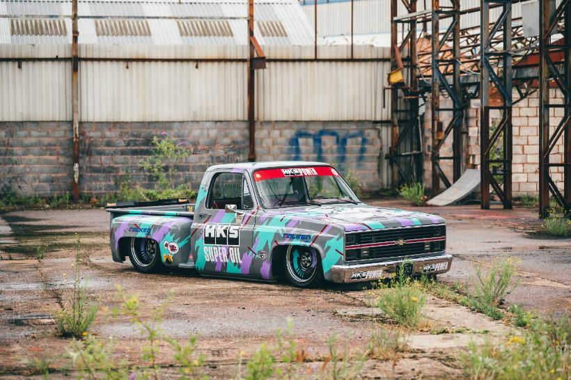 Custom slammed Chevrolet C10 pickup truck with HKS Super Oil 90s style livery parked outside an industrial warehouse, shot by 24Seven Photography.