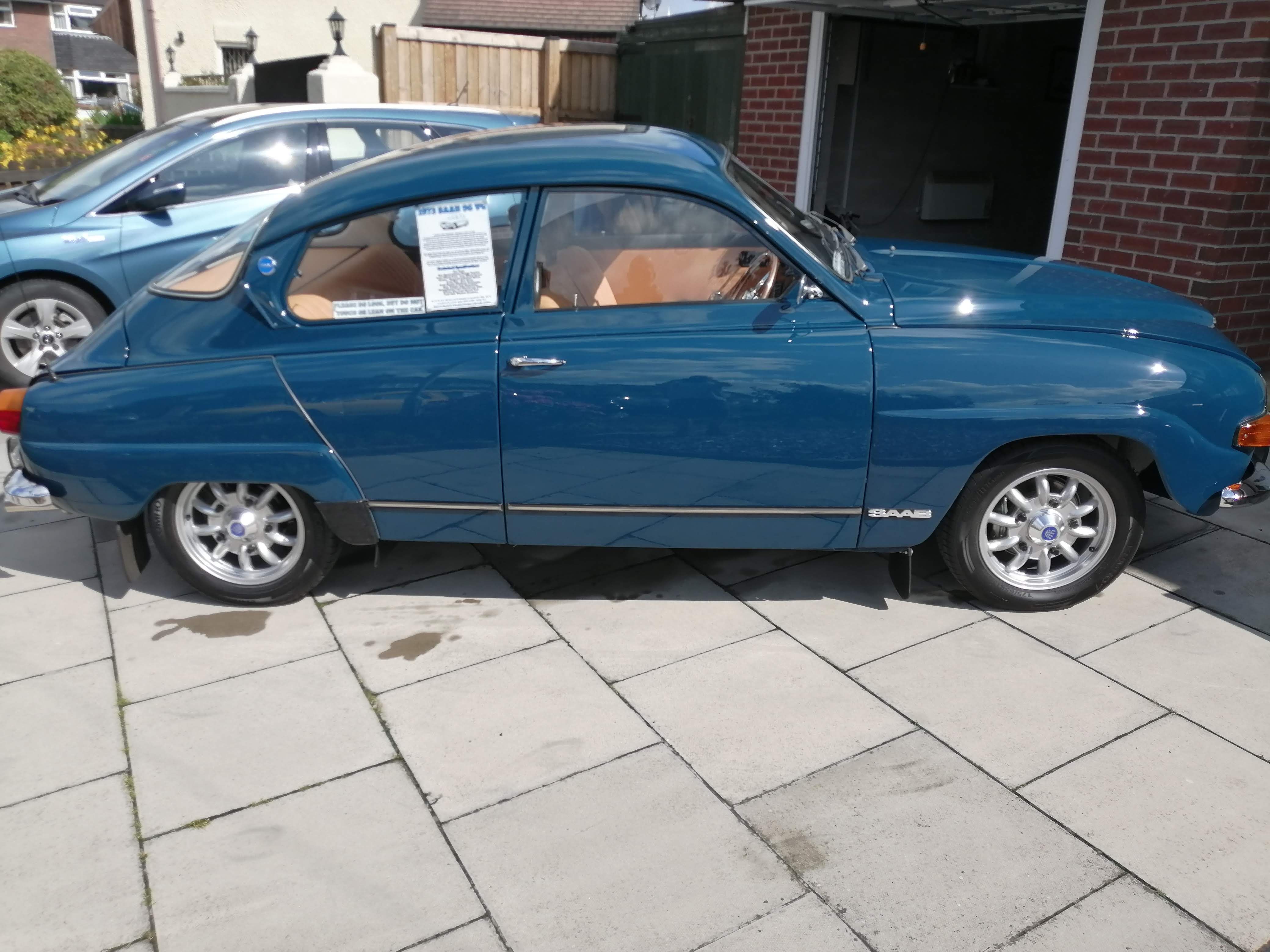 A classic teal blue Saab 96 car, owned by Christopher Boffey, parked on a paved driveway with its bonnet open, showing the tan interior.