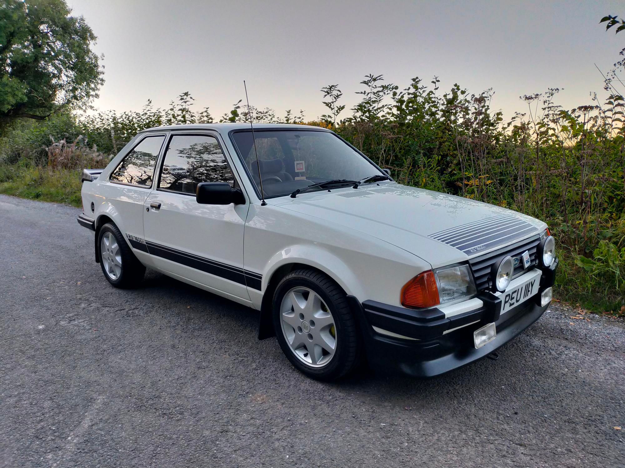 A white Ford Escort RS Turbo (Mk3) with black graphics, owned by Christopher Frost, with period-correct alloy wheels, parked on a rural road at sunset.