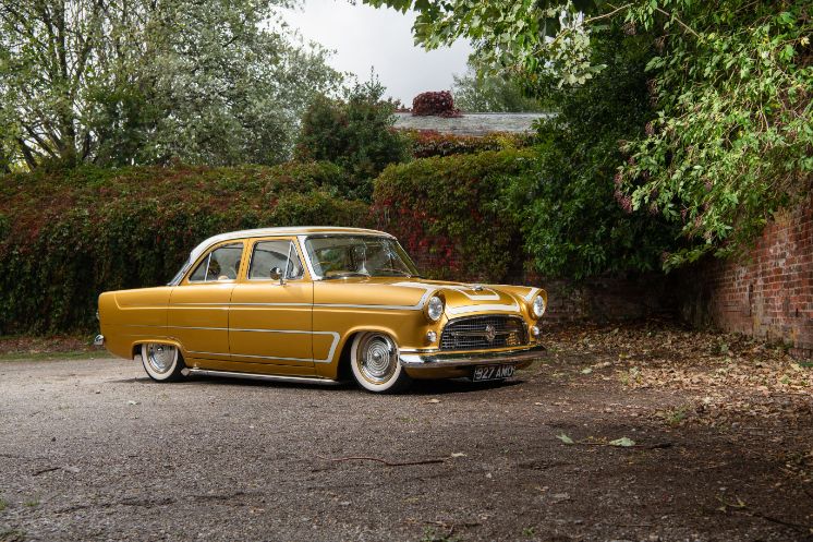 Lowrider custom Ford Consul in metallic gold paint with white pinstriping parked on gravel by a brick wall, shot by Ade Brannan.