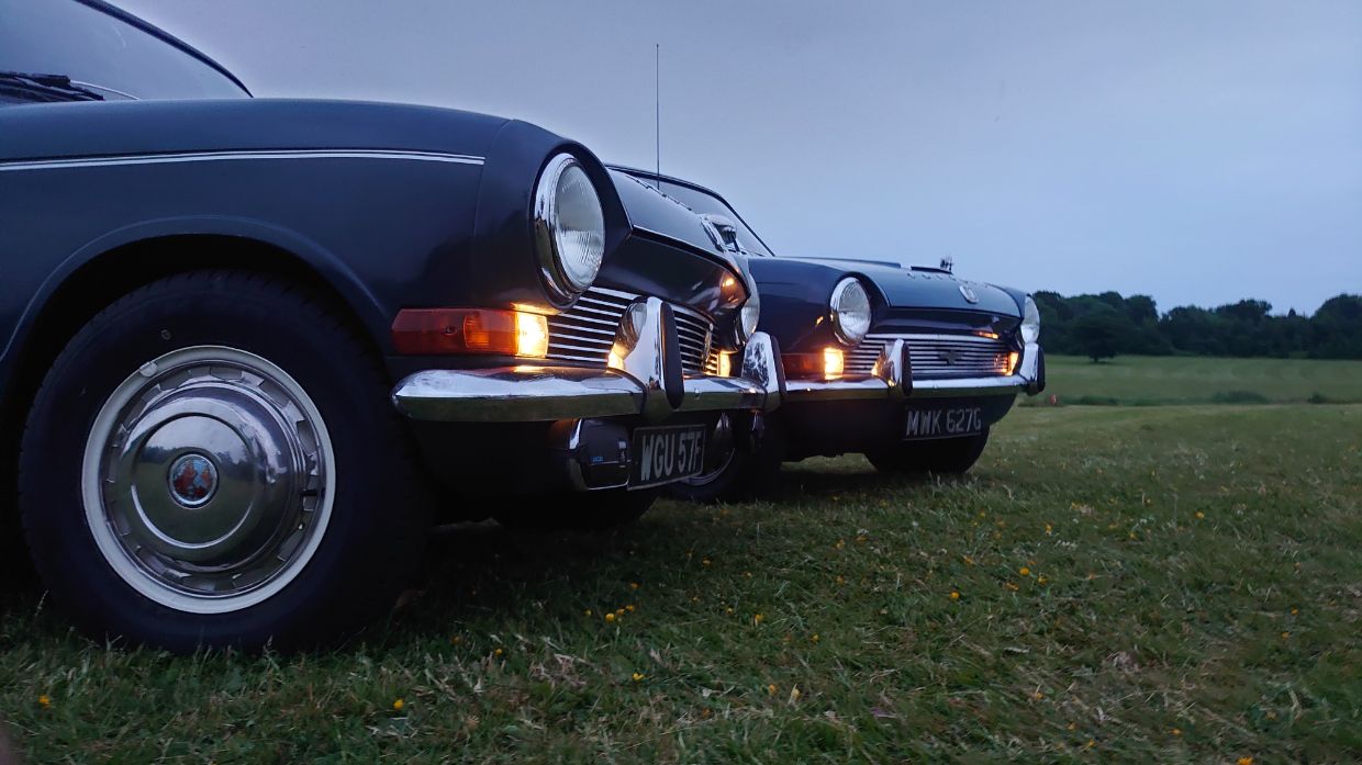 Two dark blue vintage Triumph 1300 saloons parked side-by-side on a grassy field at dusk.