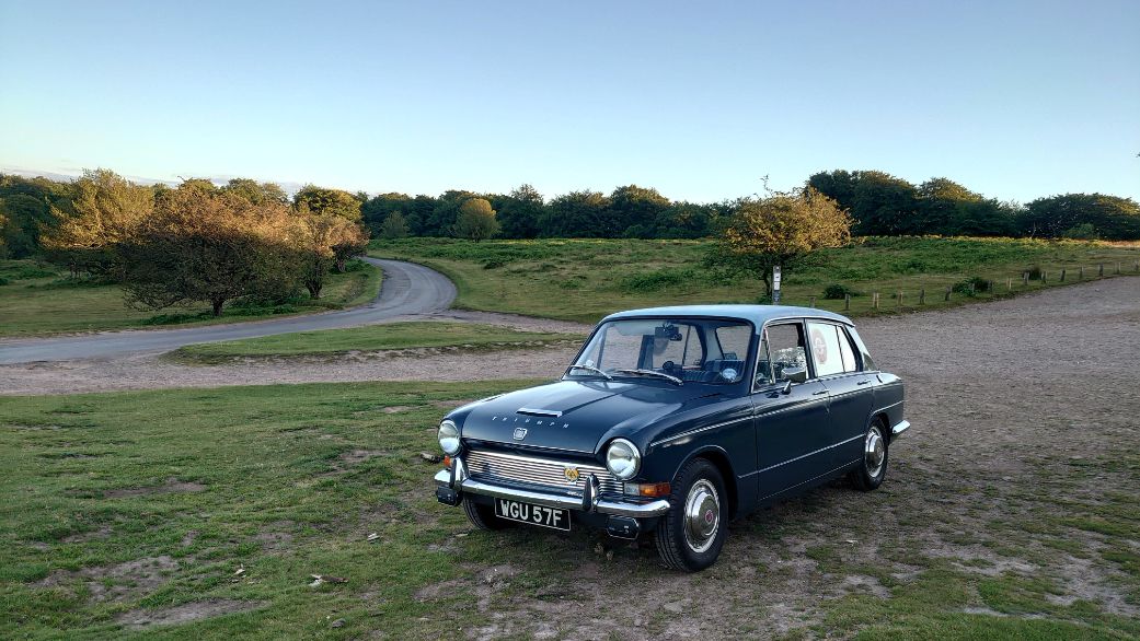Vintage dark blue Triumph 1300 saloon parked on a grassy hill, overlooking a winding road and countryside.