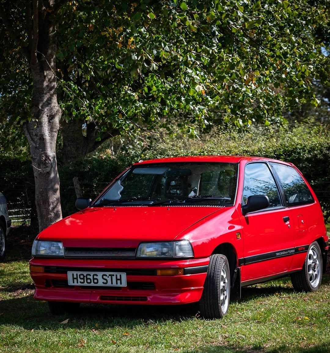A red turbocharged Daihatsu Charade GTti hot hatch, owned by Darren Adamson, with a black trim and aftermarket wheels, parked under a tree in a grassy area.