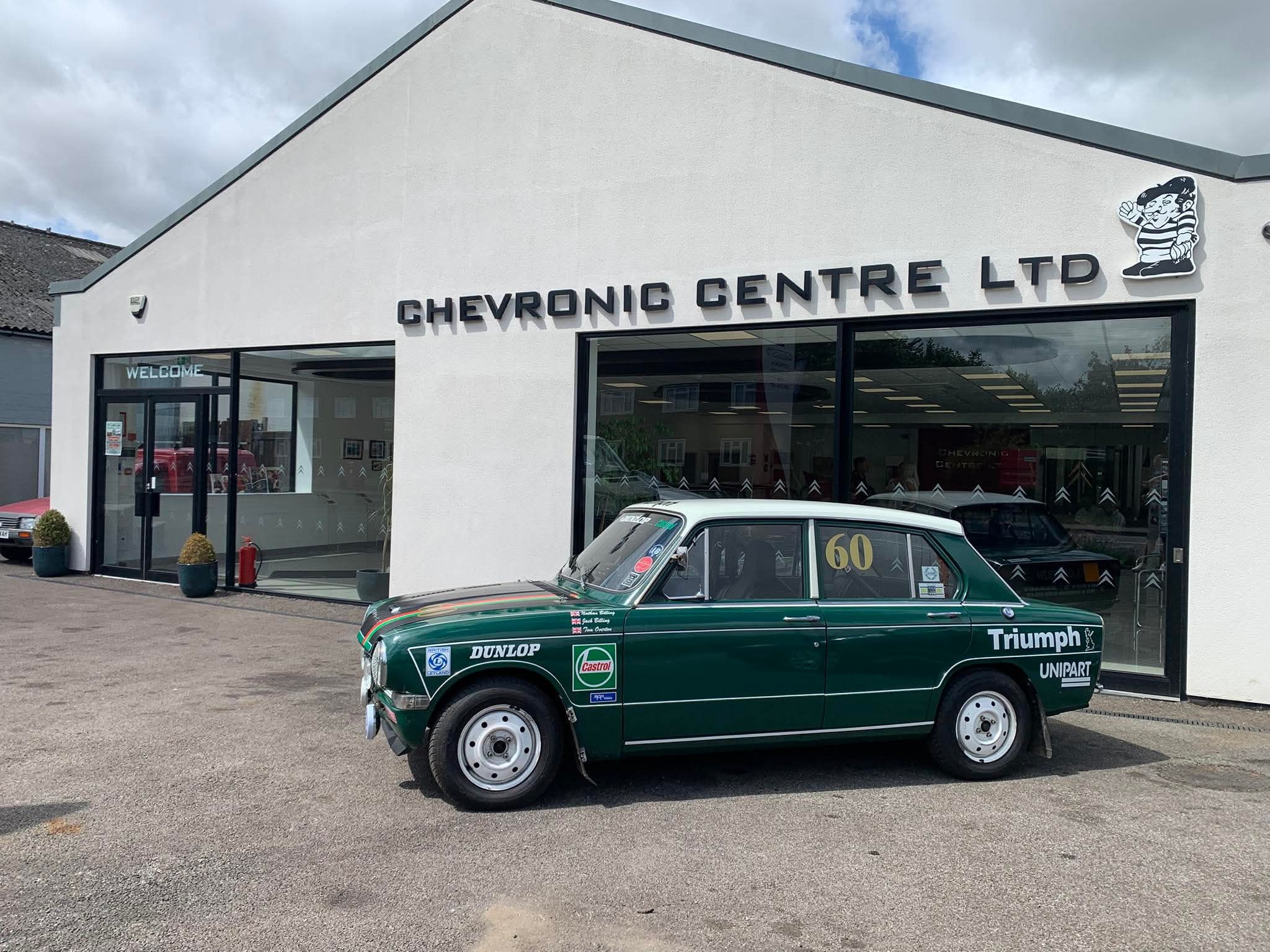 Vintage green and white Triumph 1300 rally car with competition livery parked outside Chevronic Centre Ltd, a classic car specialist.
