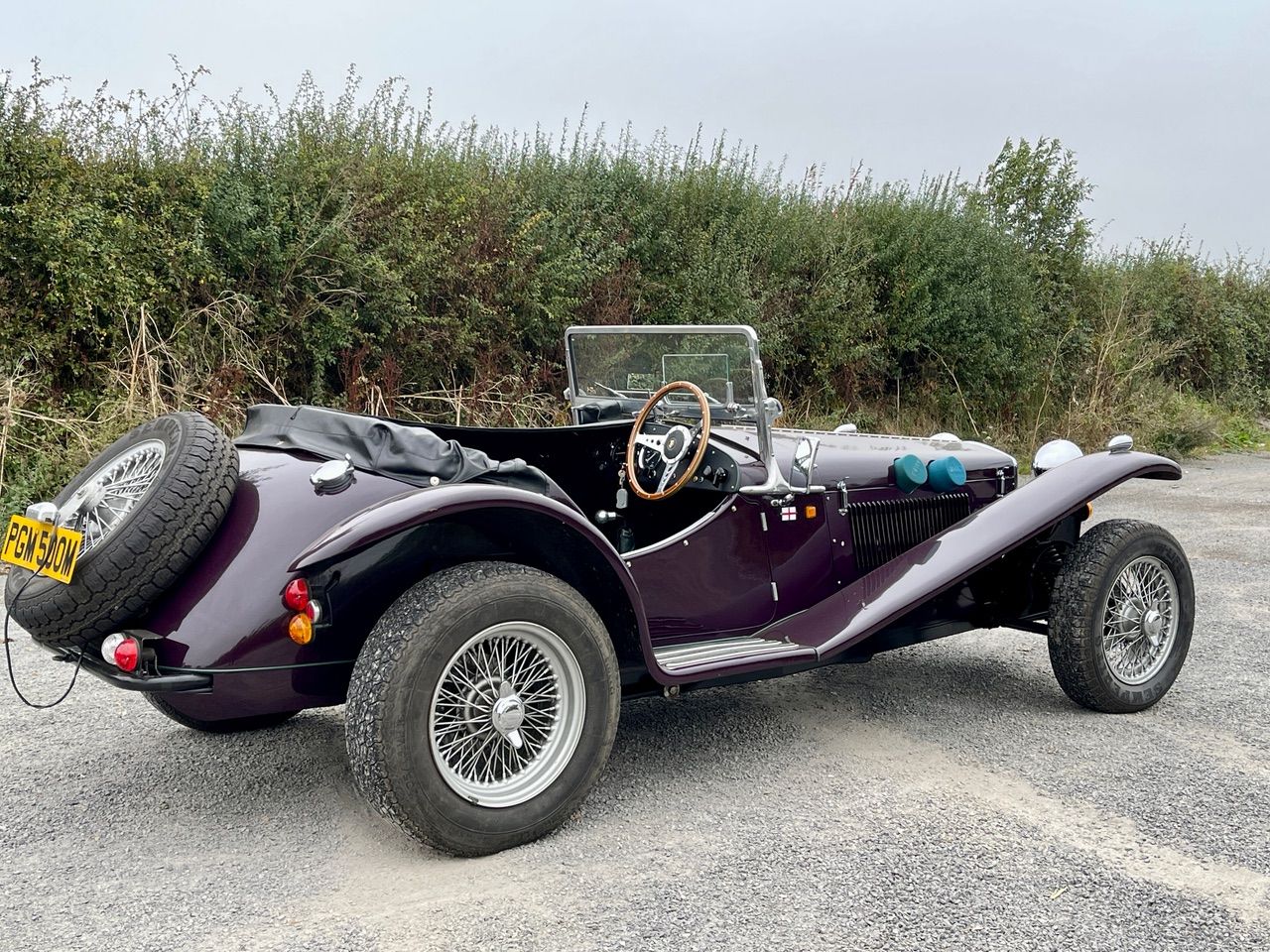 Vintage purple classic sports car with wire wheels parked on a cobbled driveway in a historic village setting.