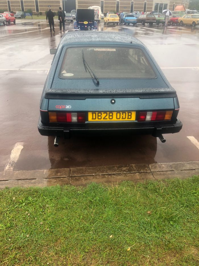 A classic dark blue Ford Capri 280, license plate "D828 ODB", is shown from the rear in a wet parking lot, with a line of other vintage cars visible in the distance.