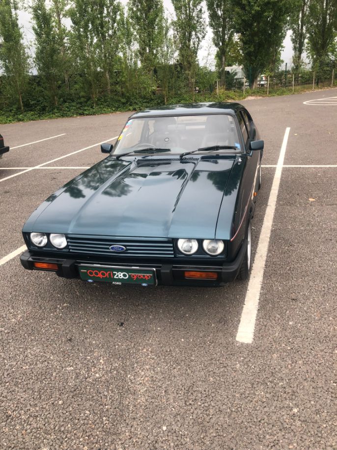 A pristine dark green Ford Capri 280 parked in a lot, with a "capri 280 group" front license plate and a line of tall trees in the background.