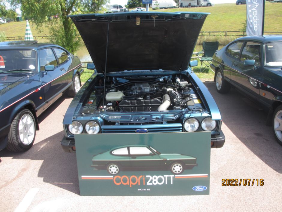 A dark green Ford Capri 280TT with its hood open, showcasing the engine at an outdoor car show, with two other classic Ford Capris visible on either side and a display board for the "Capri 280TT" in the foreground.