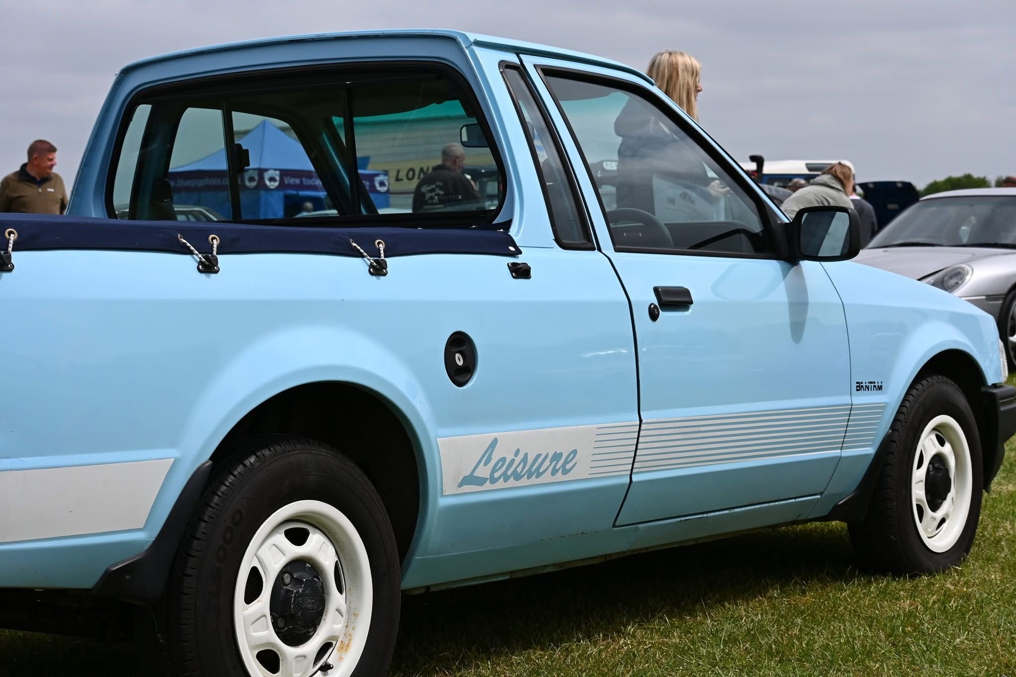 A classic light blue Ford Escort Bantam "Leisure" pickup truck, with a blue bed cover and white wheels, photographed from the side at an outdoor car show.