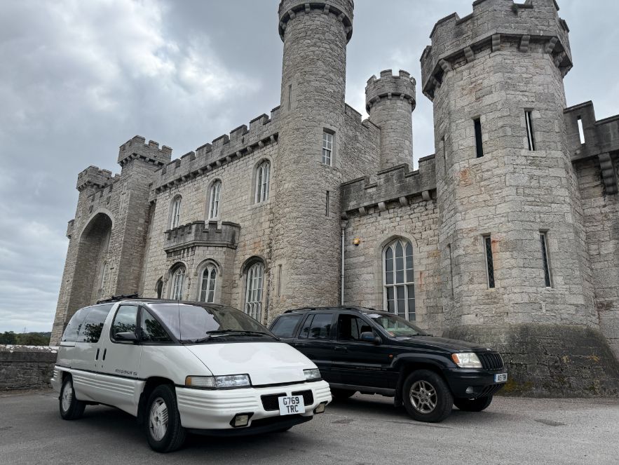 A white Pontiac Trans Sport minivan and a black Jeep Grand Cherokee parked in front of a stone castle.