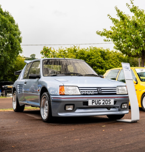 A rare, light blue Peugeot 205 T16 rally replica (Group B styling), owned by John Hemming, on display with custom wide arches and wheels at an outdoor car event.