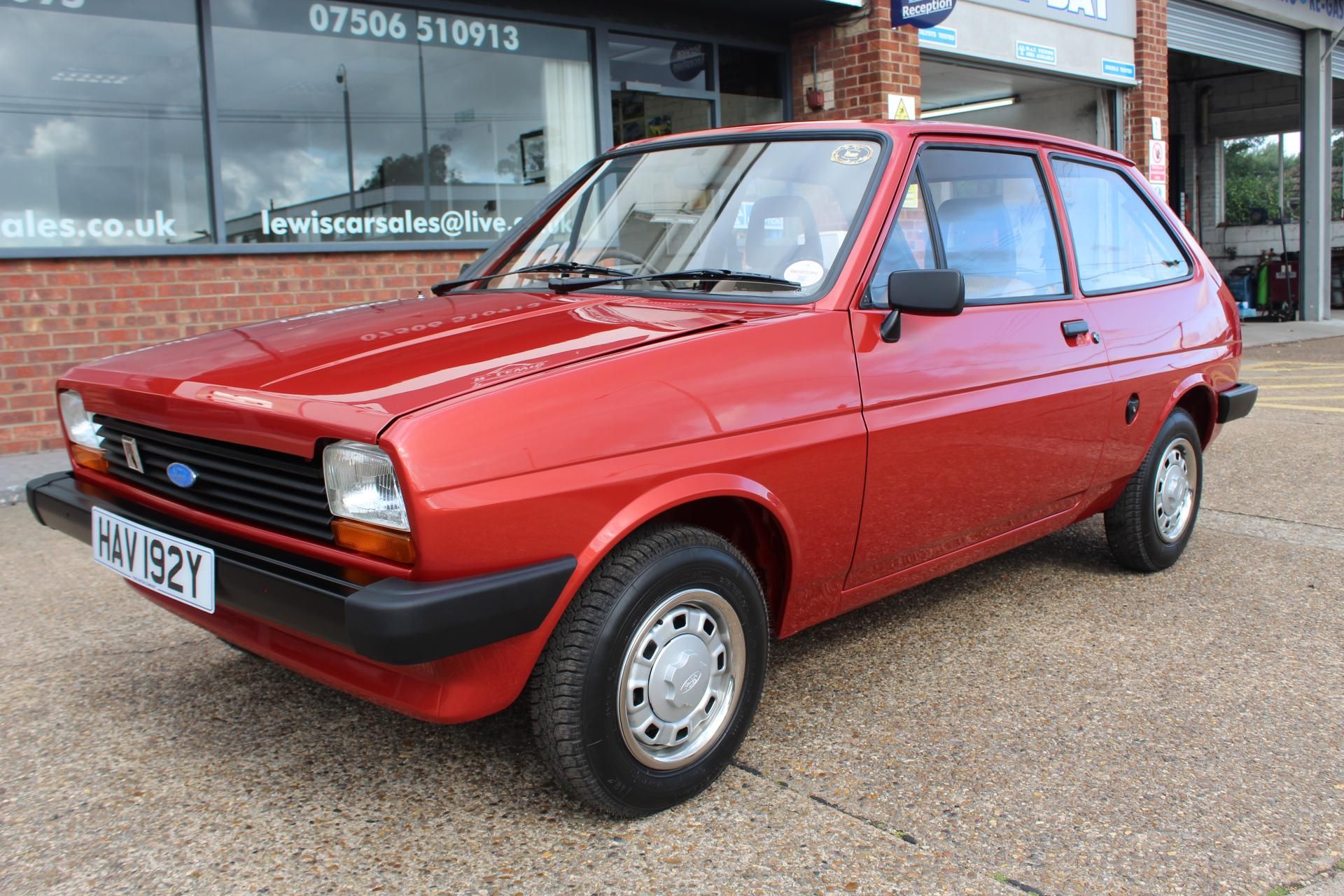 A restored bright red Ford Fiesta Mark I hatchback, owned by Jordan Fenton, with original silver hubcaps, parked outside a brick building.