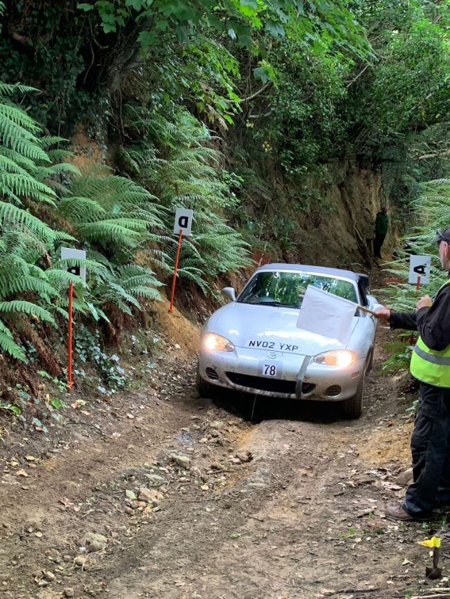 Silver Mazda MX-5 rally car navigating a muddy trial course at Knowle Lane, during a classic car trials event, with a marshal guiding.