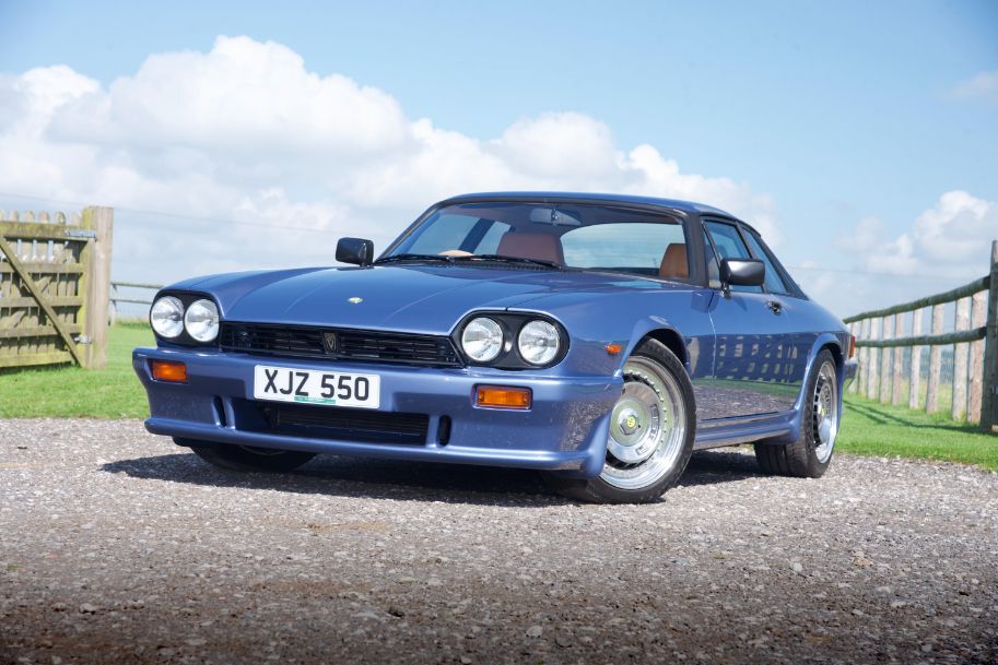 Front view of a vintage blue Lister XJ-S parked on a gravel driveway with a field behind it.
